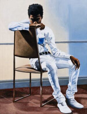An adolescent boy sits in a chair with his chin tucked under his arm wearing an all white outfit